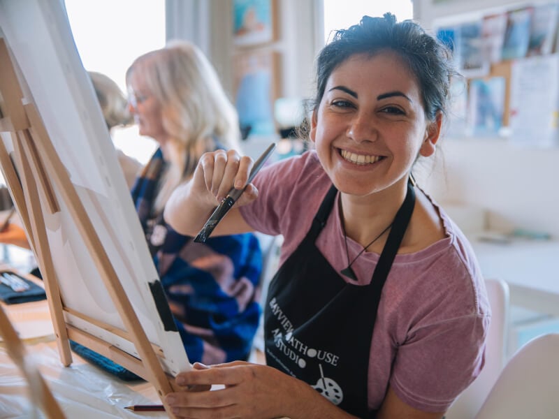 A woman smiles as she holds a brush to a canvas at a painting class